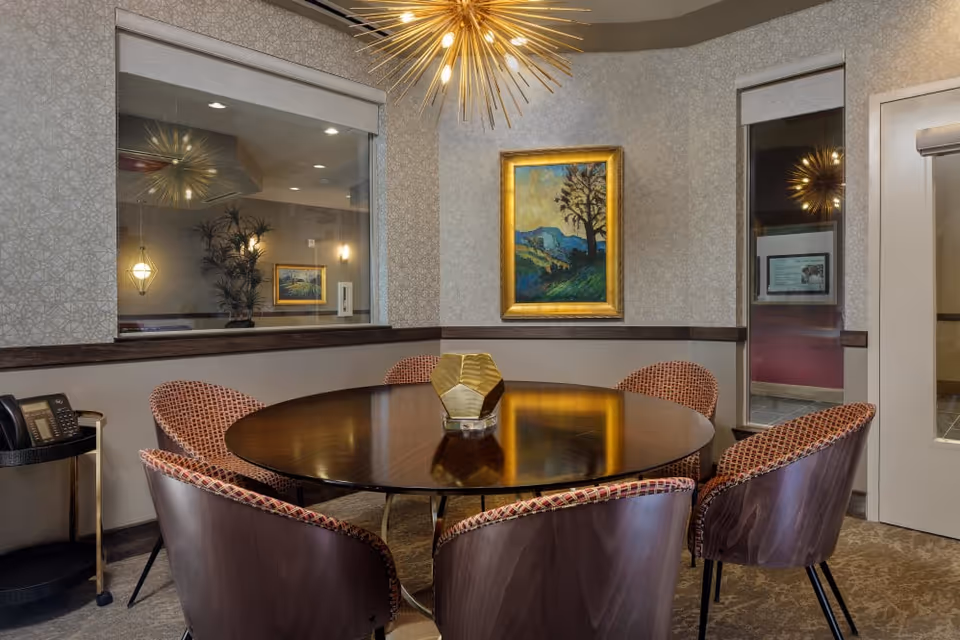 A small meeting or dining room with a round wooden table surrounded by six upholstered chairs with patterned fabric. A modern gold geometric centerpiece sits on the table. The walls have a light patterned wallpaper and a framed landscape painting. A large window looks into another room, and a modern gold starburst chandelier hangs from the ceiling. A phone is placed on a small black and gold side table in the corner.
