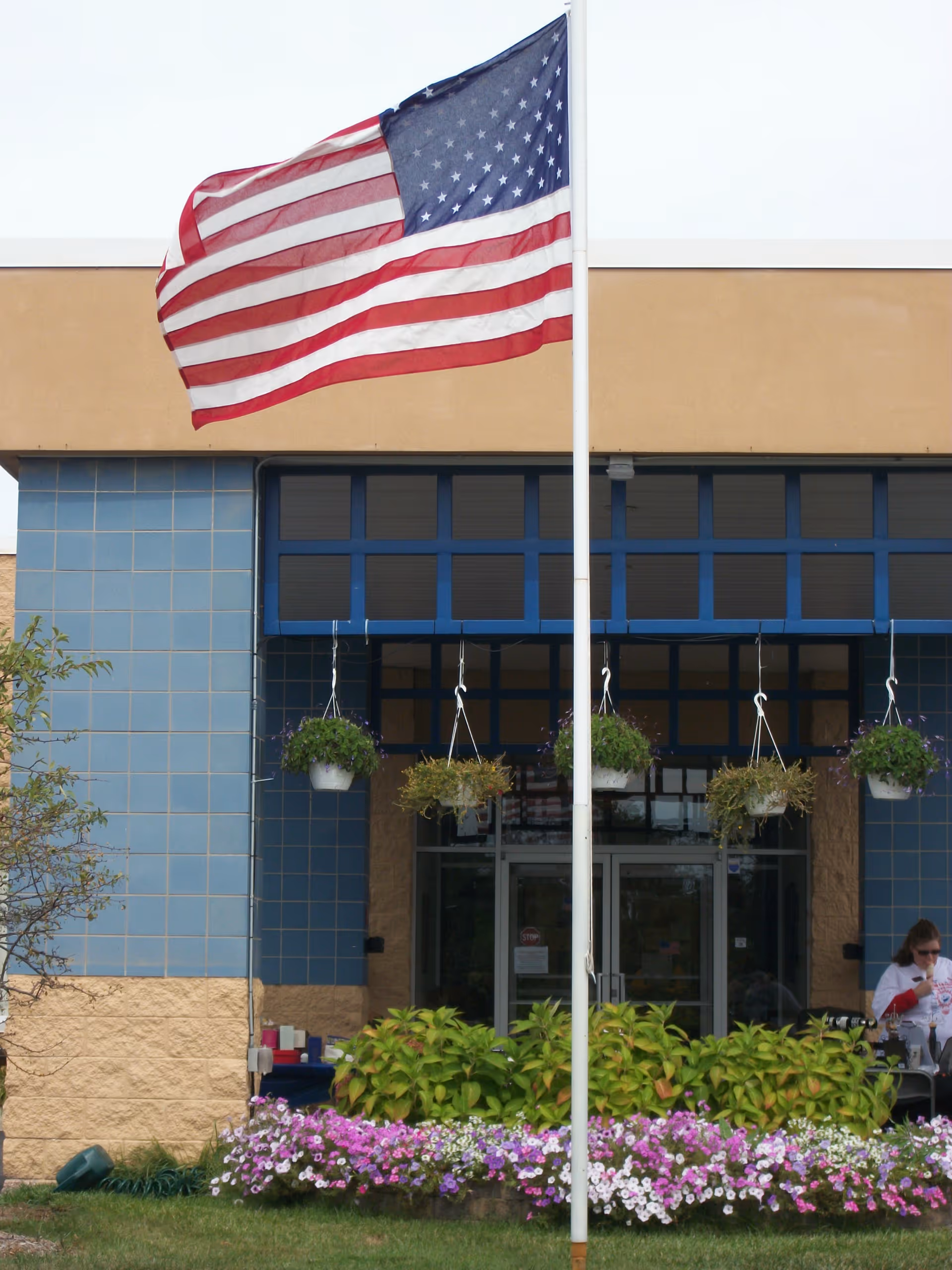 An American flag flies on a pole outside a building entrance with hanging flower baskets and a colorful flowerbed.