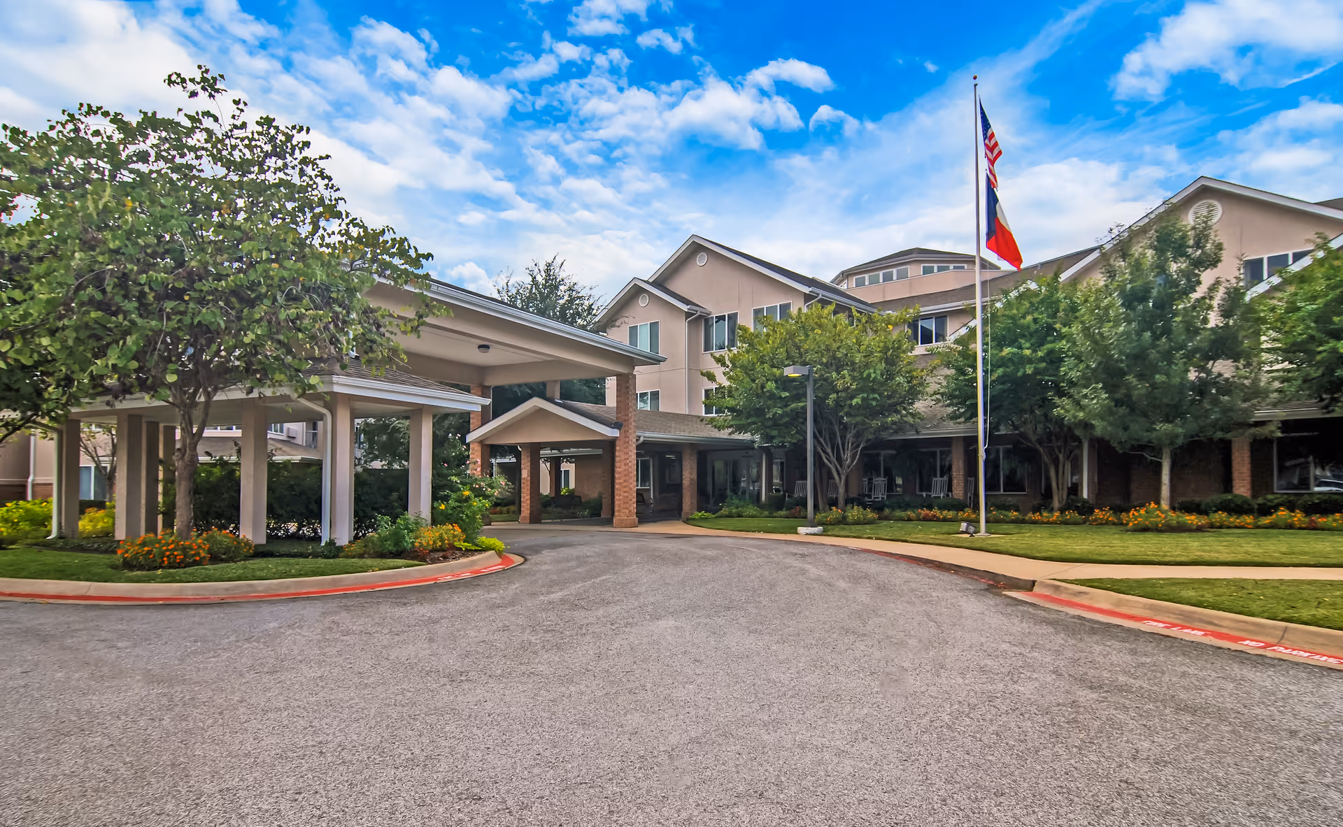 Entrance and front façade of a multi-story senior living facility with a covered drop-off, flagpoles, landscaping, and a circular driveway.