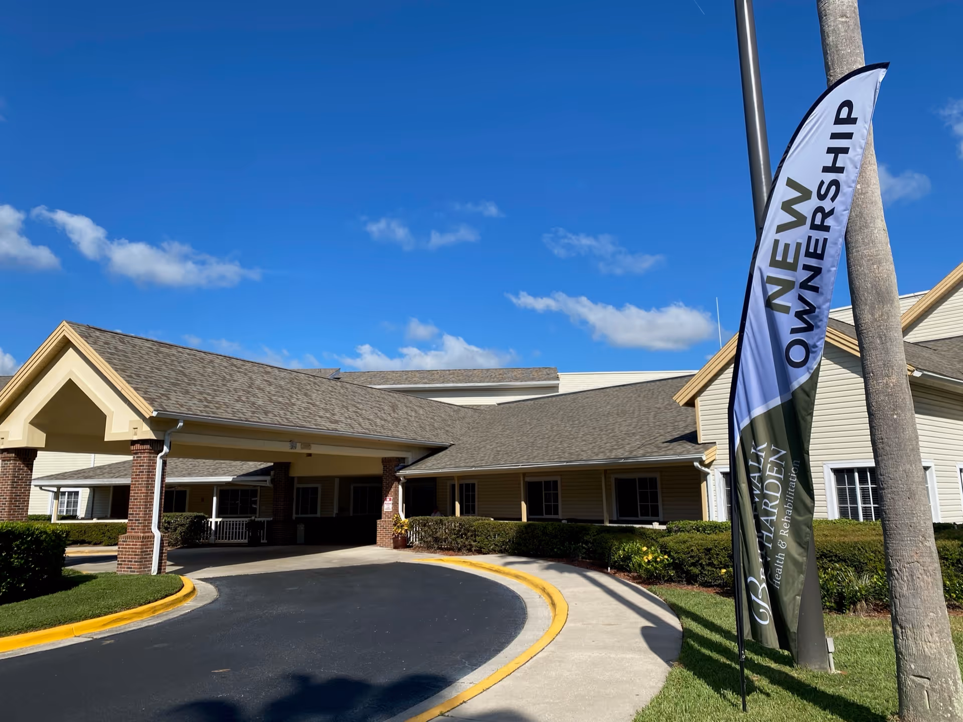 Exterior view of Bridgewalk on Harden facility showing the entrance with a covered drop-off area supported by brick columns. A flag near a palm tree reads 'NEW OWNERSHIP'. The building has beige siding and a shingled roof under a clear blue sky.