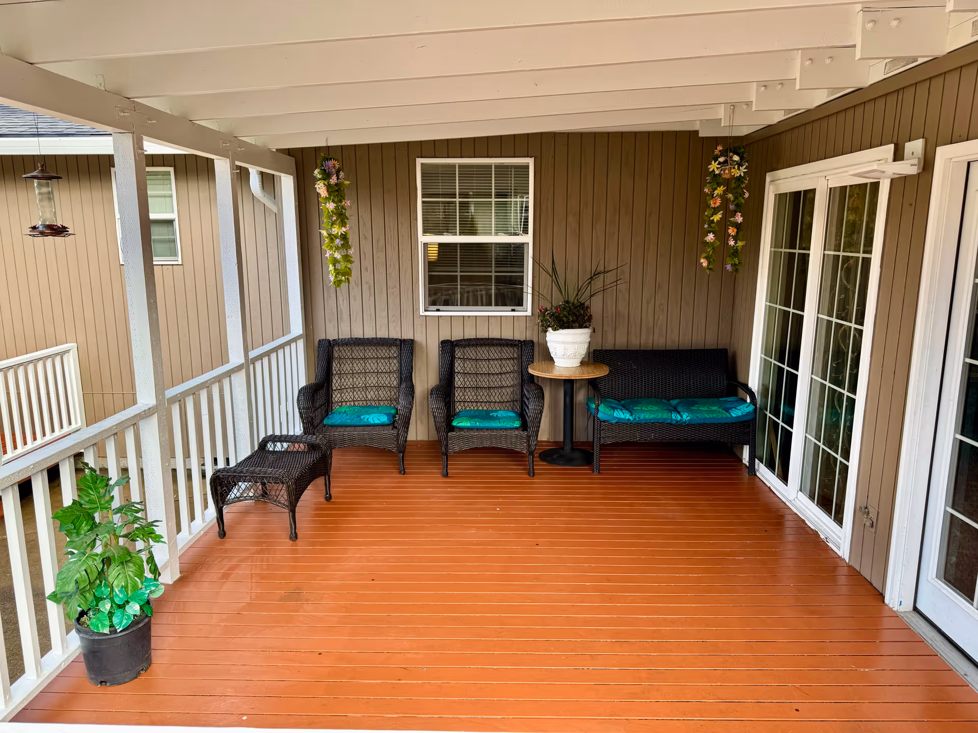 Covered outdoor porch area with brown wooden floor and walls, featuring two wicker chairs and a wicker loveseat with teal cushions, a small round table with a potted plant, hanging flower decorations, a potted plant on the floor, and white railing along the edge.