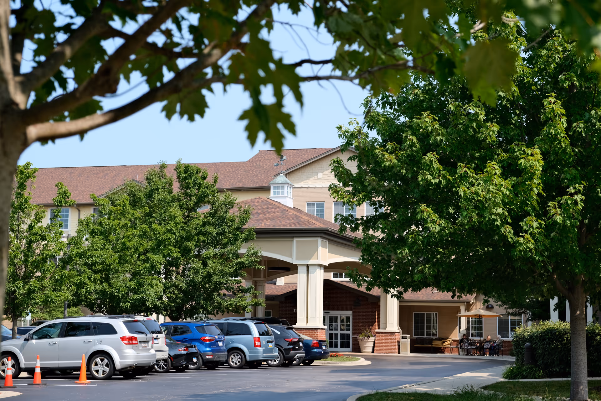 Front entrance of a senior living building framed by trees with parked cars in the driveway.