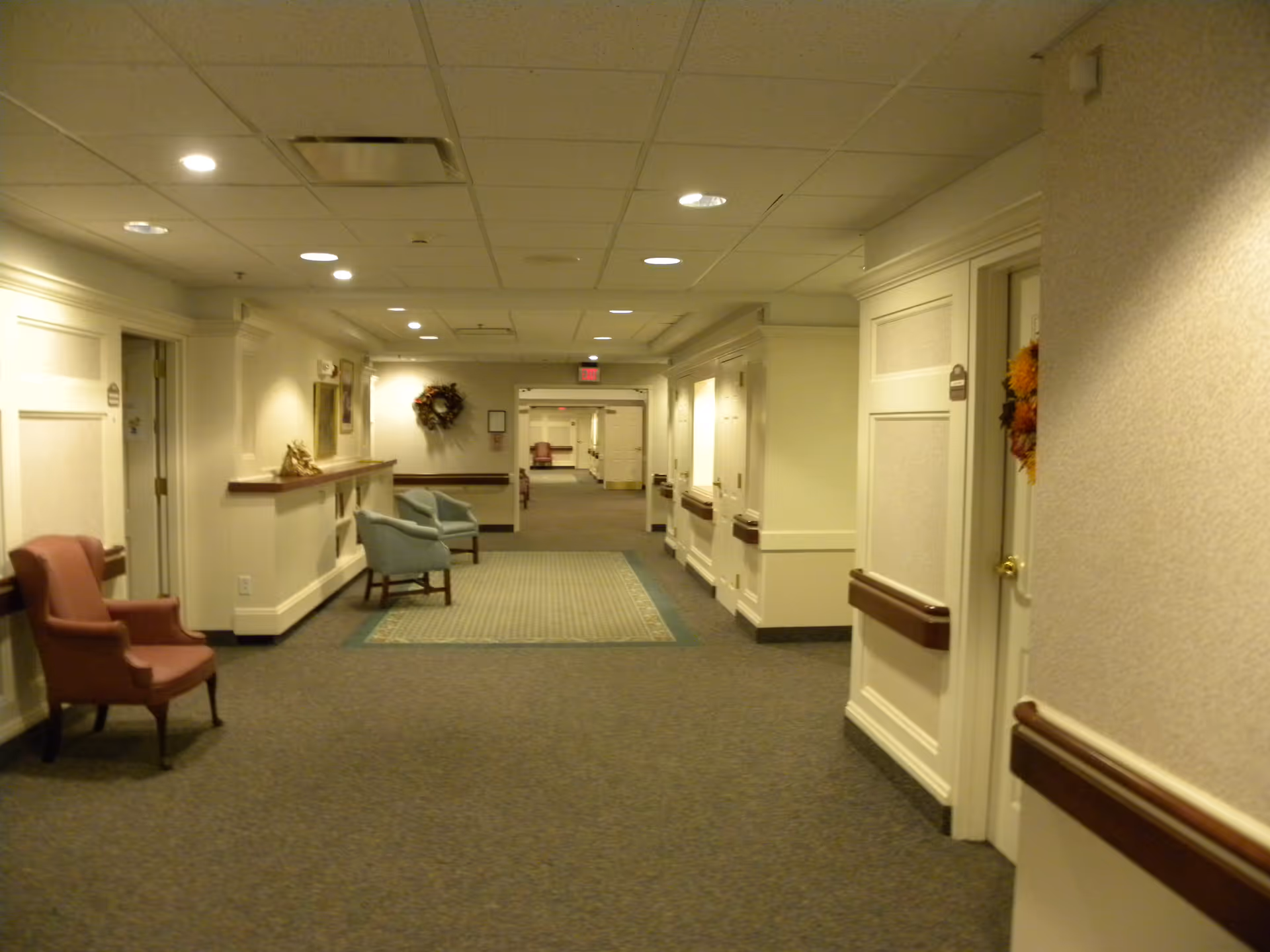 A well-lit hallway in a senior living facility with beige walls and carpeted floor. Several chairs are placed along the walls, and there are handrails on both sides. The hallway extends into the distance with doors on either side and a wreath hanging on the wall at the far end.