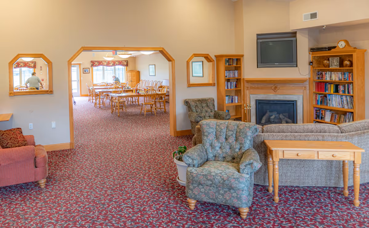 Cozy senior living lounge with fireplace, chairs, bookshelves, and a dining room beyond.