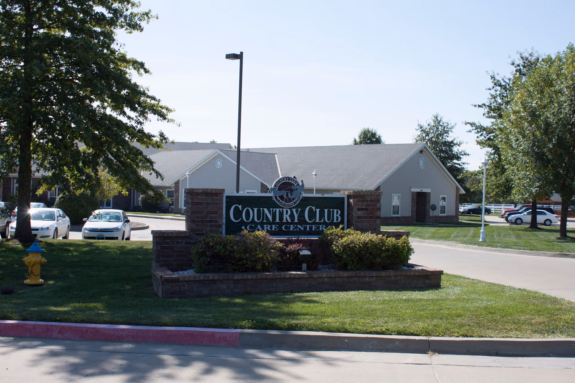 Entrance sign reading 'Country Club Care Center' in front of a single-story care building with parked cars and landscaping.