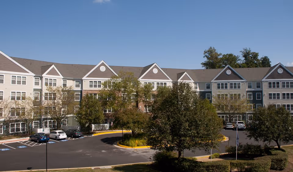 Exterior view of a multi-story senior living facility building with a parking lot in front, several cars parked, and trees surrounding the area under a clear blue sky.