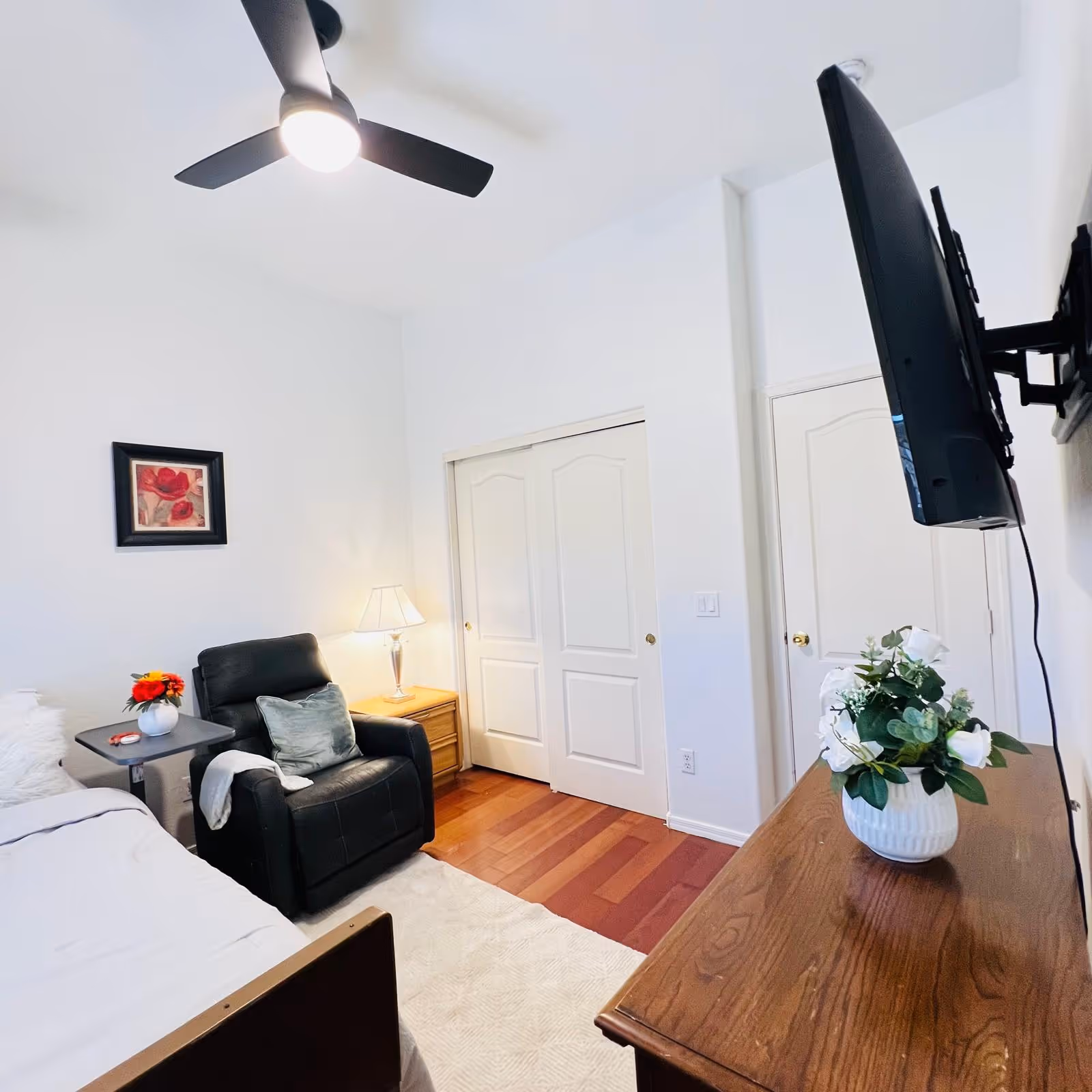 A tidy bedroom with a bed, leather chair, dresser, mounted TV, and ceiling fan.