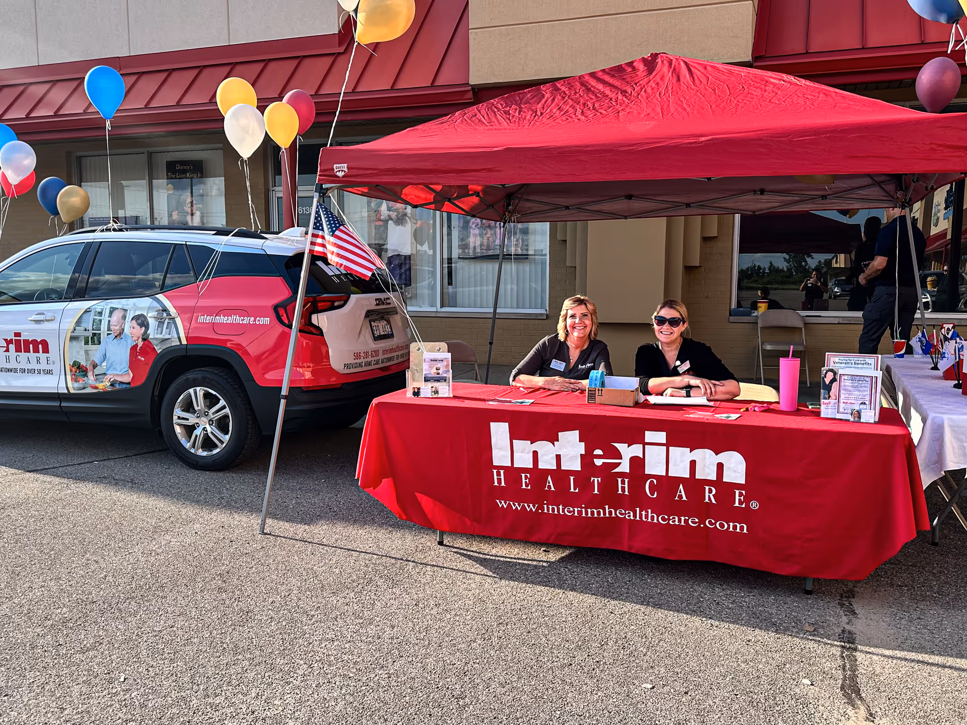 Outdoor promotional booth with two staff under a red canopy beside a branded vehicle.