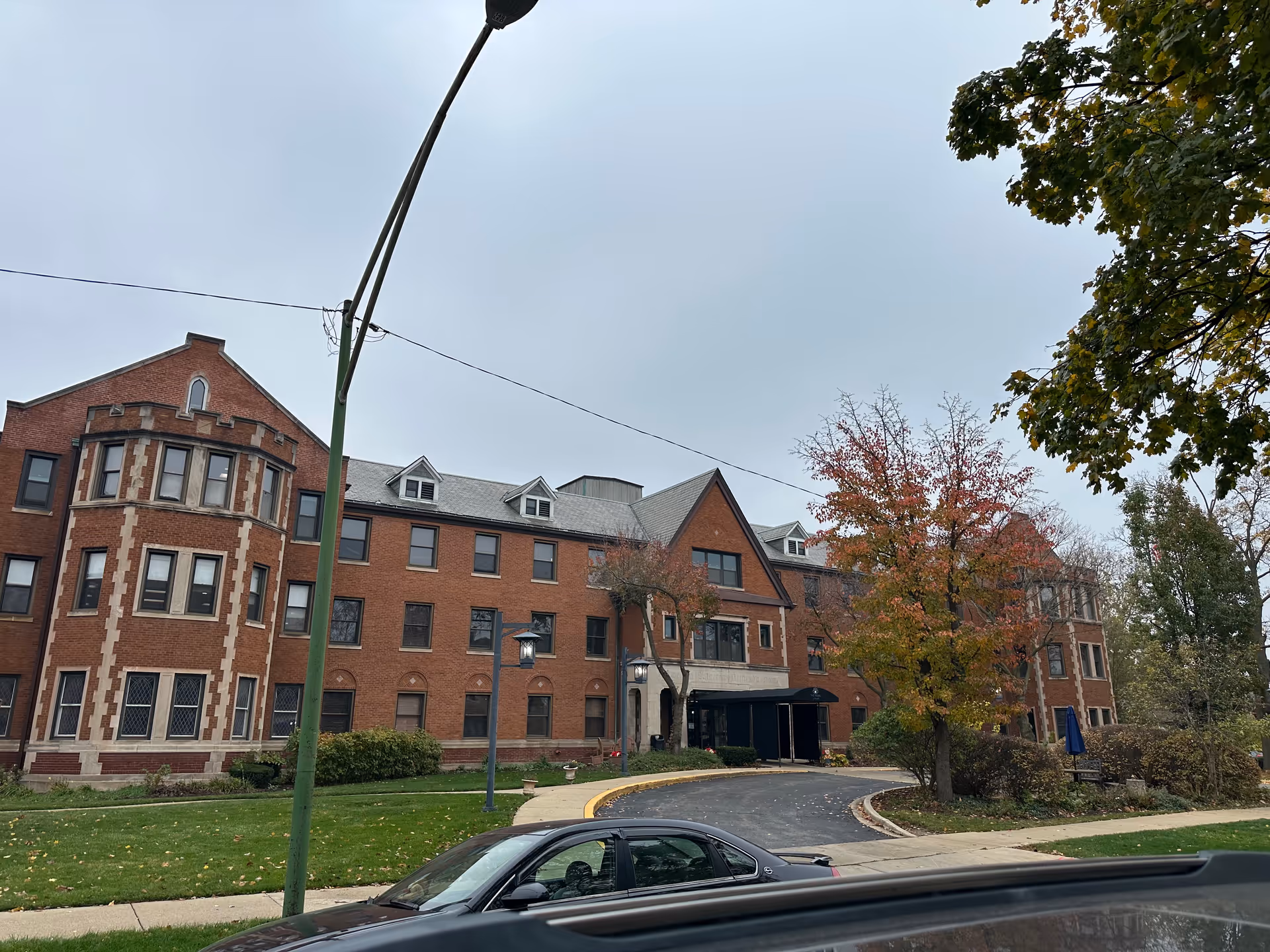Exterior view of a large three-story brick building with multiple windows and a peaked roof. The building is surrounded by green grass, trees with autumn foliage, and a curved driveway with a black car parked nearby. The sky is overcast.