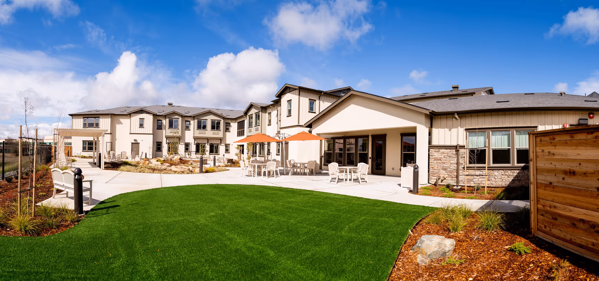 Outdoor courtyard area of CountryHouse Residence featuring a well-maintained green lawn, several white patio tables with orange umbrellas, white chairs, and benches. The building surrounding the courtyard is two stories with beige and stone exterior walls under a blue sky with scattered clouds.