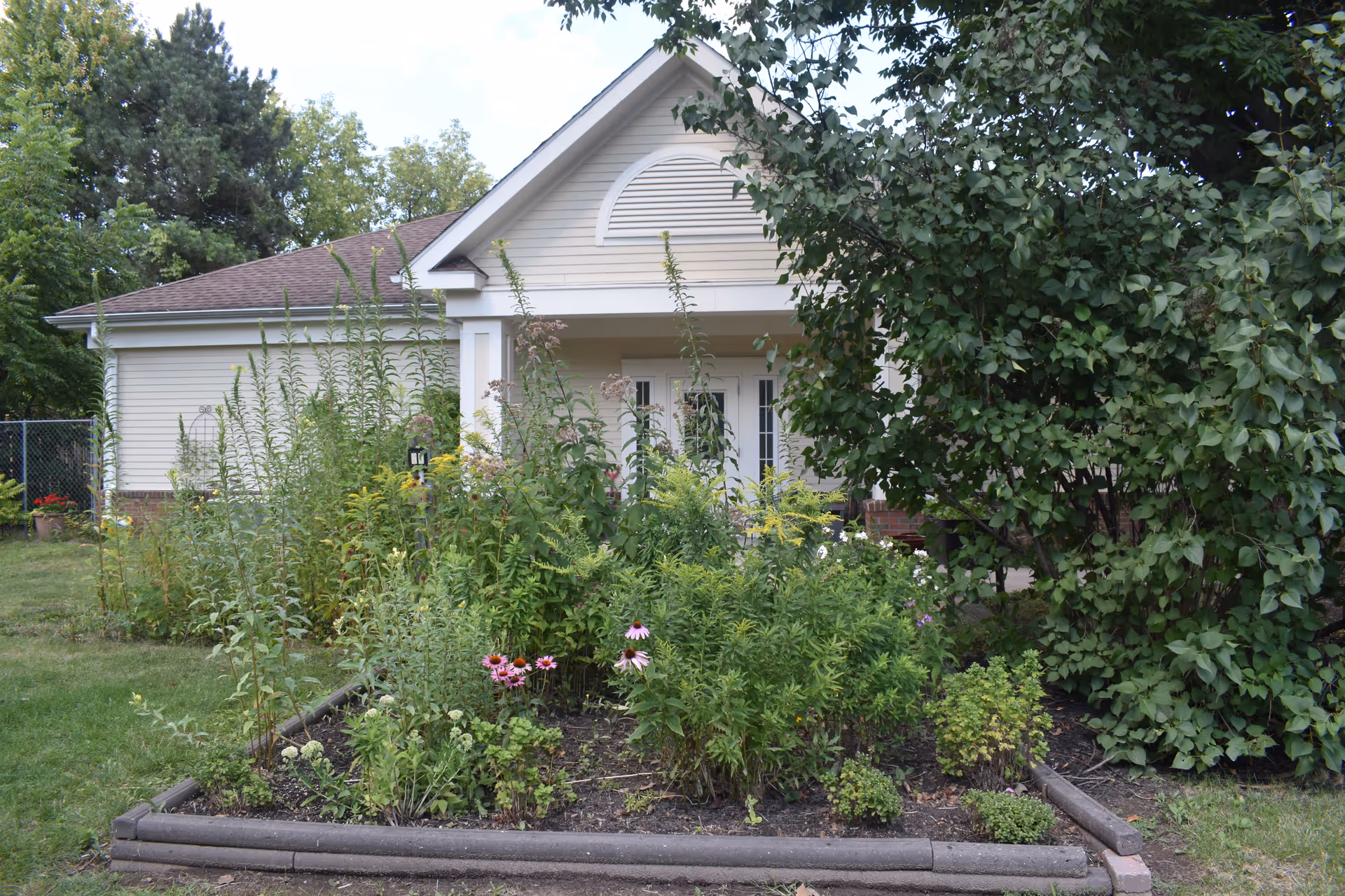 Front exterior view of a single-story building with beige siding and a gabled roof, partially obscured by a garden with various green plants and flowers in the foreground.