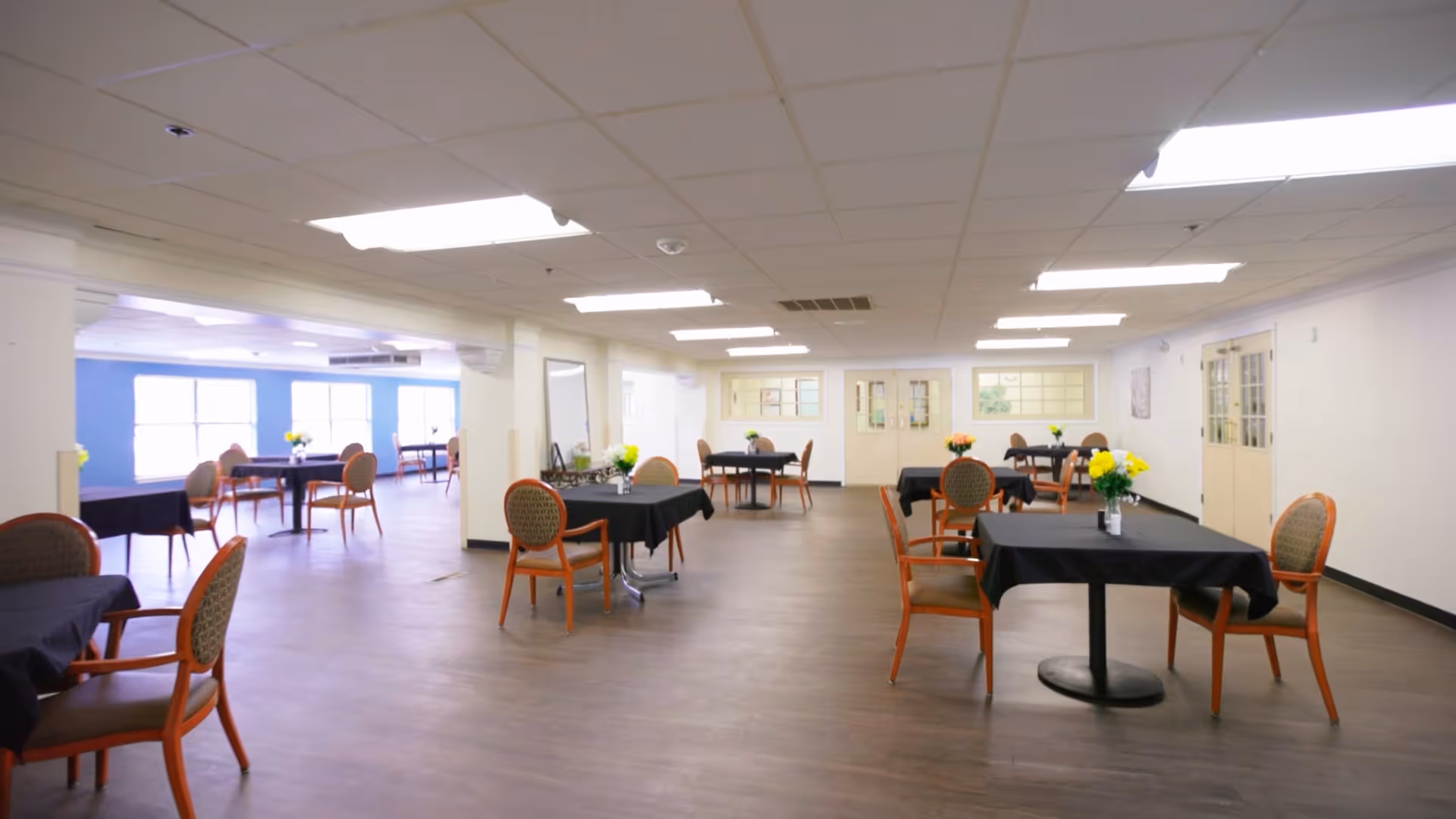 Spacious senior dining/activity room with round tables covered in black cloths, chairs, and small floral centerpieces.