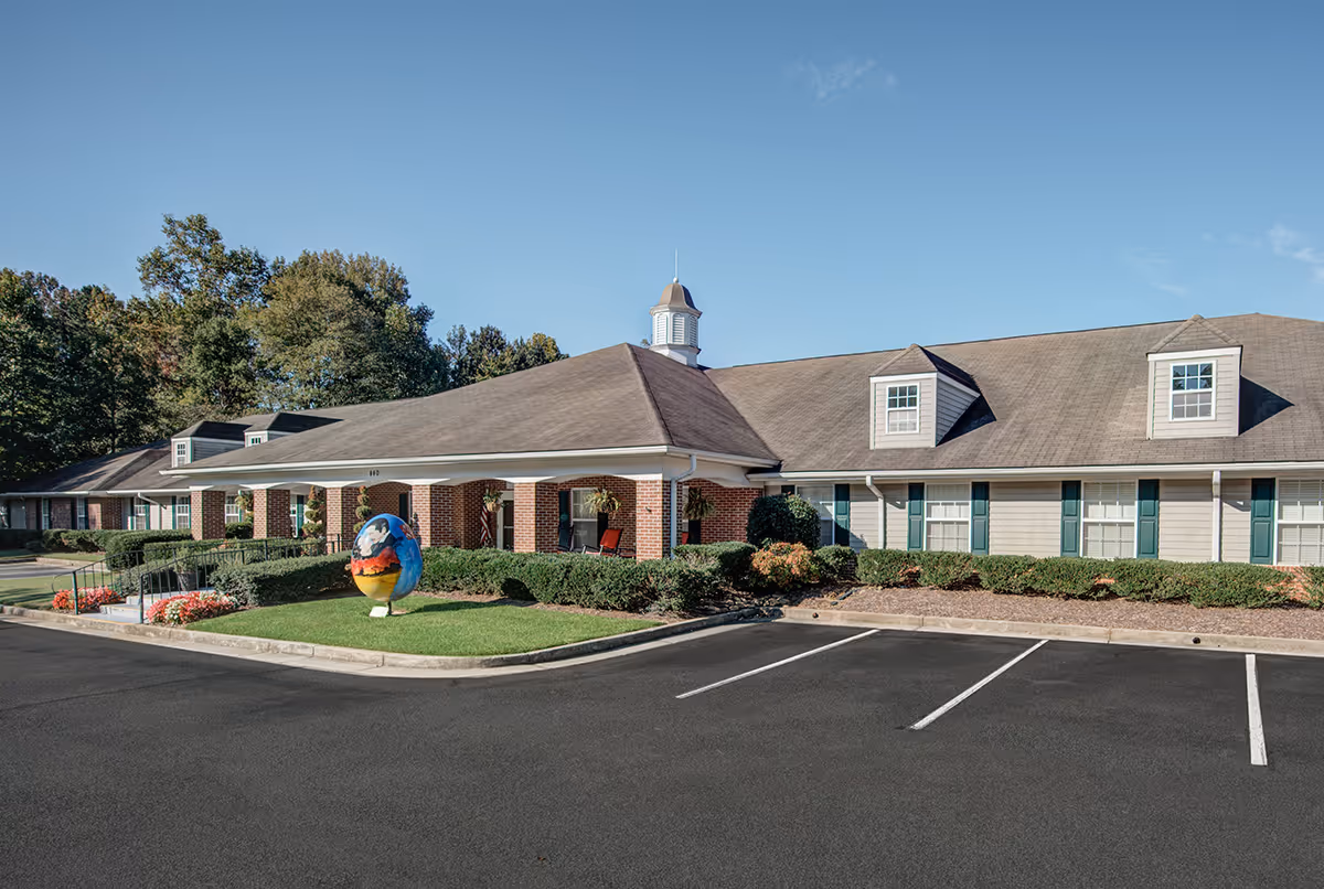 Exterior view of a single-story senior living facility building with a pitched roof, dormer windows, and a covered entrance. There is a small landscaped area with bushes and flowers, and a colorful decorative globe sculpture on the lawn near the entrance. The parking lot in front has marked parking spaces and the sky is clear and blue.