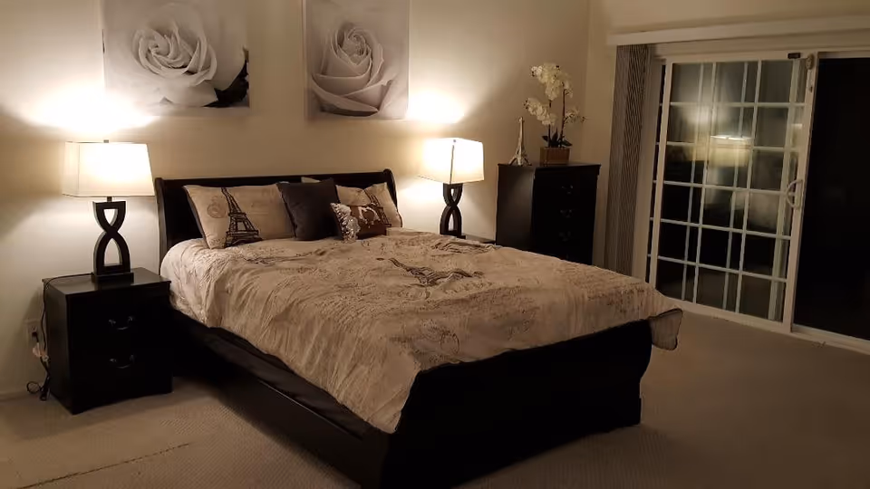 A cozy bedroom with a large bed covered in beige bedding featuring Eiffel Tower designs. There are two matching bedside tables with modern lamps on each side of the bed. Above the bed, two framed pictures of white roses are hung on the wall. To the right, there is a dark wooden dresser with a decorative plant and a small Eiffel Tower figurine. A sliding glass door with vertical blinds is also visible, leading outside or to another room.