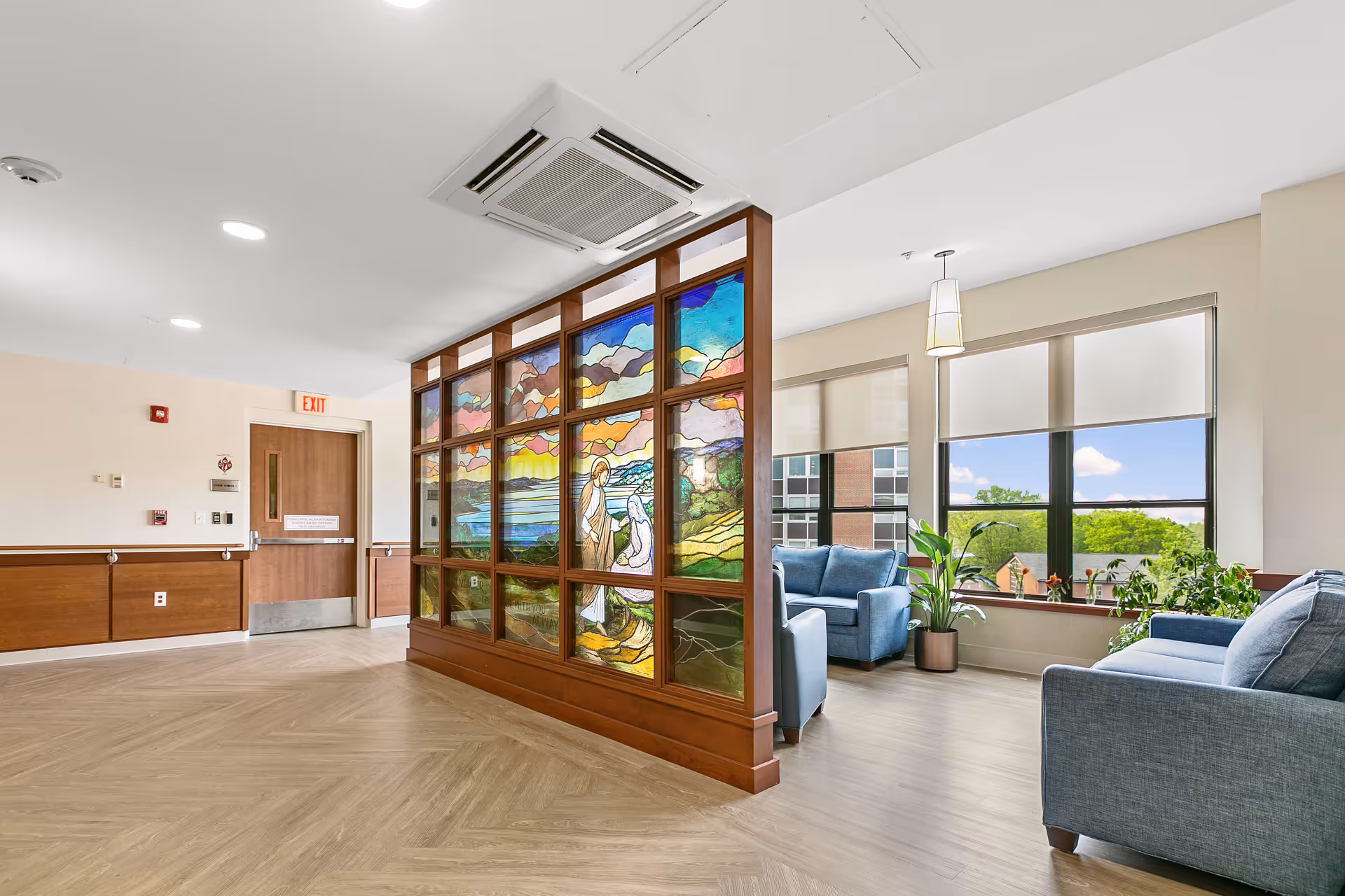 Bright senior living lounge with blue chairs, plants, and a decorative stained glass partition.