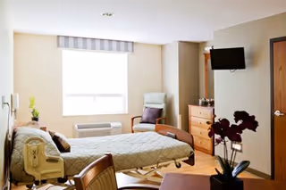 A bedroom in a senior living facility with a bed, dresser, chair, and wall TV.
