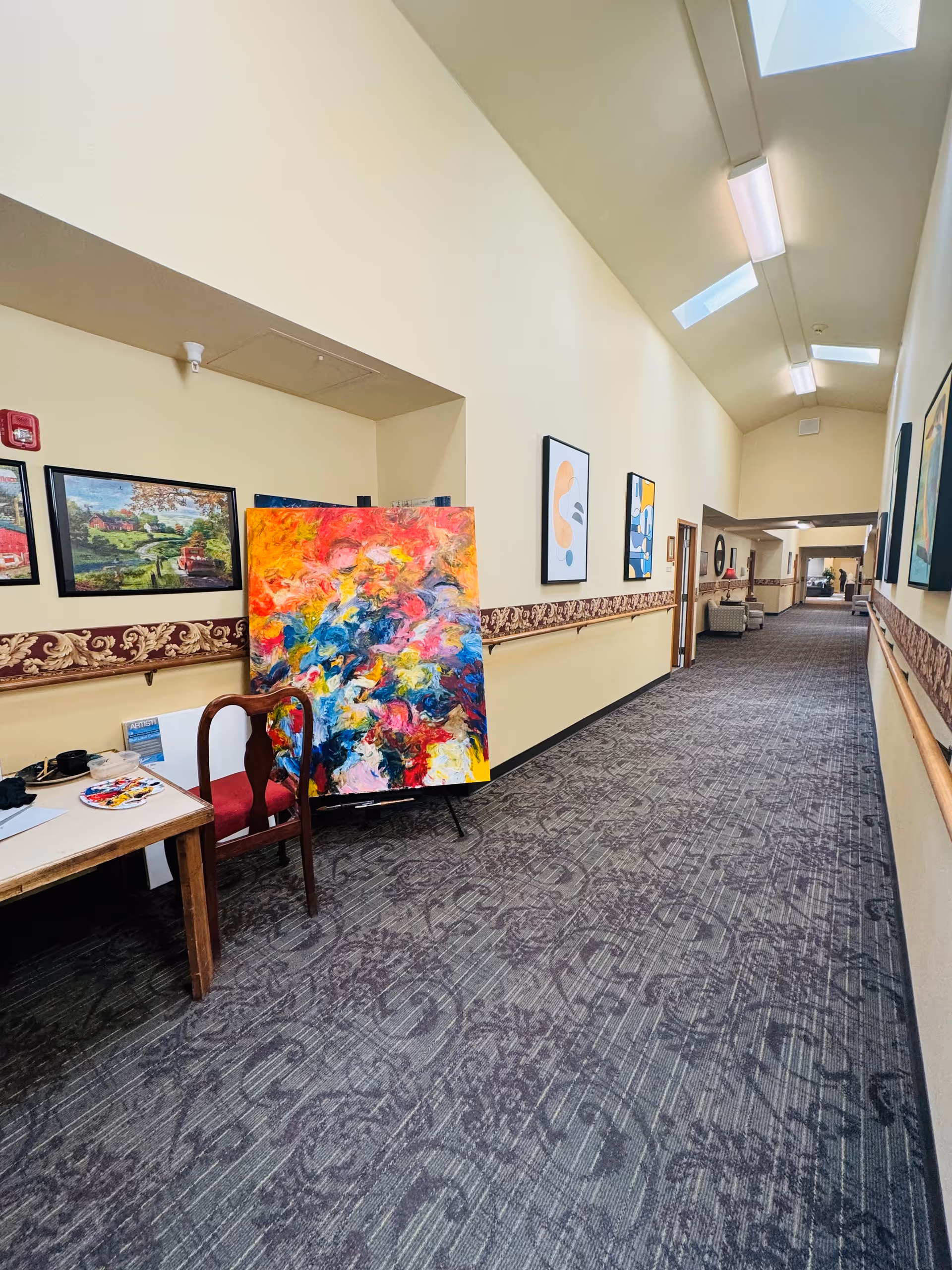 Interior hallway of a senior living facility with colorful paintings and a table.