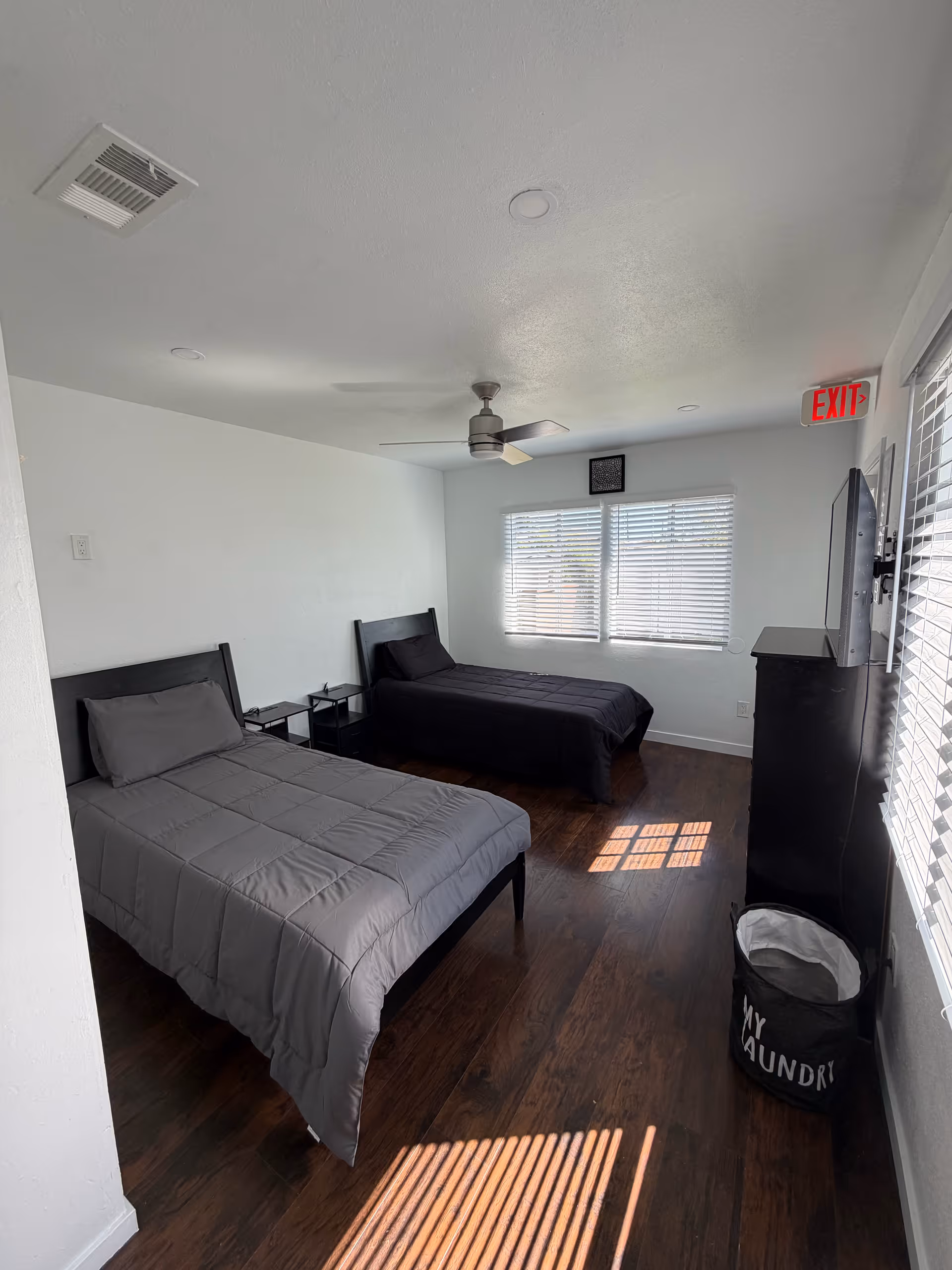Two twin beds with gray bedding in a bedroom, dark wood floor, blinds, and ceiling fan.