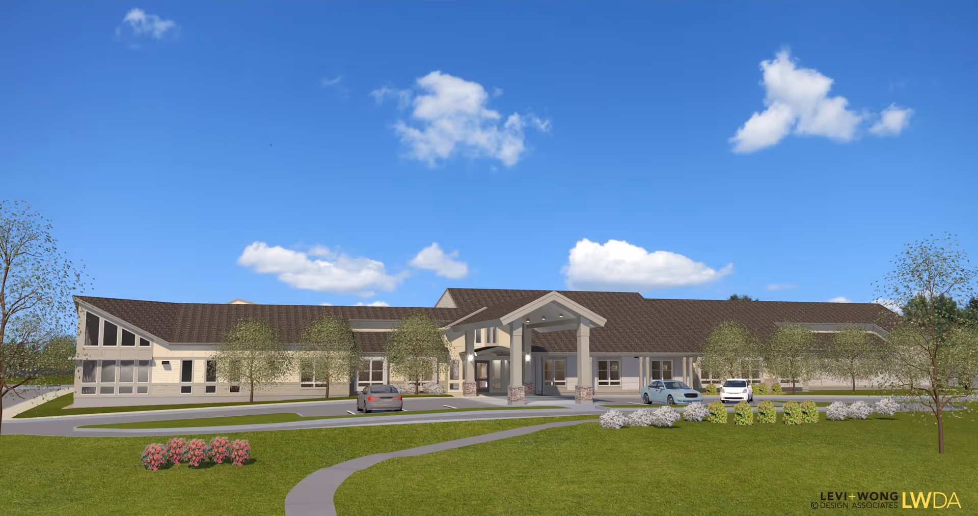 Exterior view of a single-story senior living facility building with a brown roof and beige walls under a blue sky with scattered clouds. The building has a covered entrance with columns and several windows. There are a few cars parked in front, surrounded by green lawns, trees, and landscaped bushes.