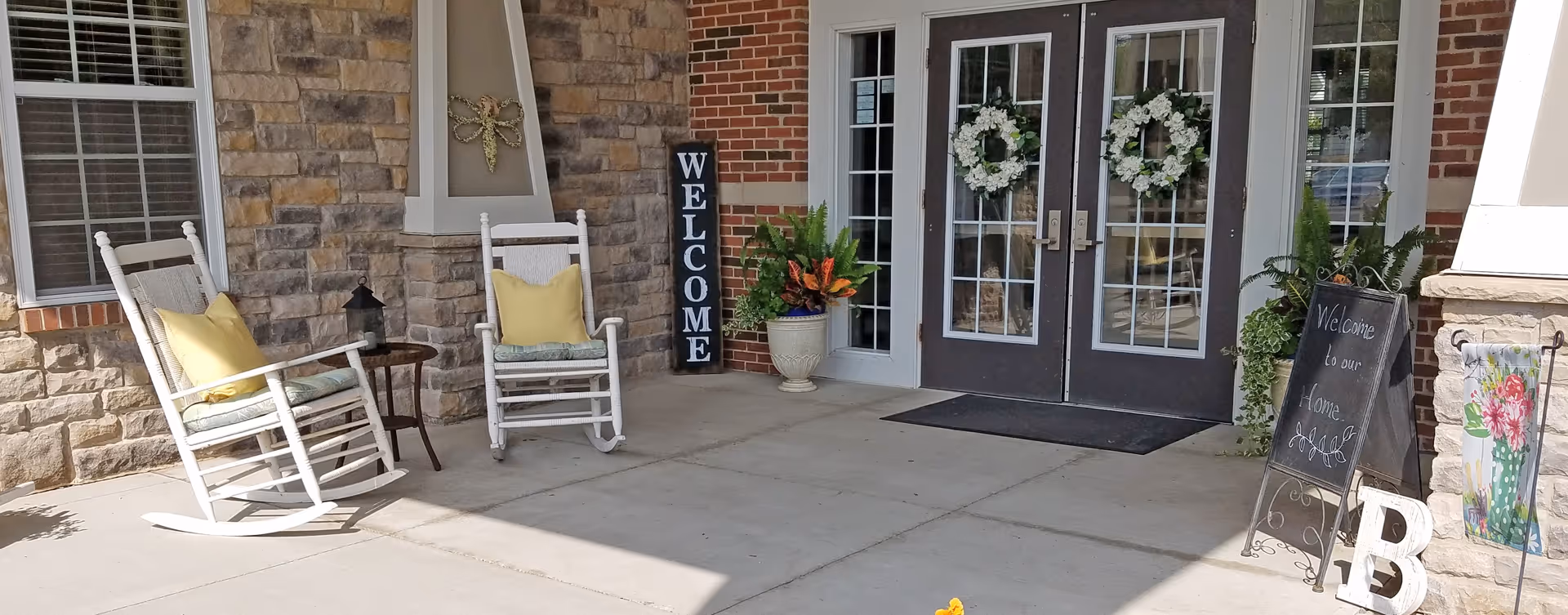 Entrance area of a facility with two white rocking chairs with yellow pillows, a small round table, a stone and brick wall, double glass doors decorated with white floral wreaths, a vertical welcome sign, potted plants, and a chalkboard sign that reads 'Welcome to our Home.'