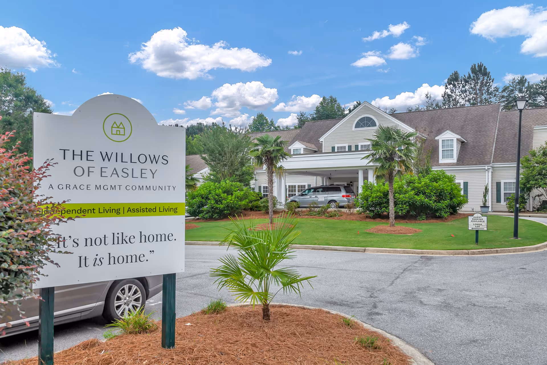 Exterior view of The Willows of Easley senior living facility with a large sign in the foreground that reads 'The Willows of Easley, A Grace MGMT Community, Independent Living | Assisted Living, It's not like home. It is home.' The building is surrounded by green landscaping, palm trees, and a driveway with a parked vehicle under a covered entrance. The sky is blue with scattered clouds.