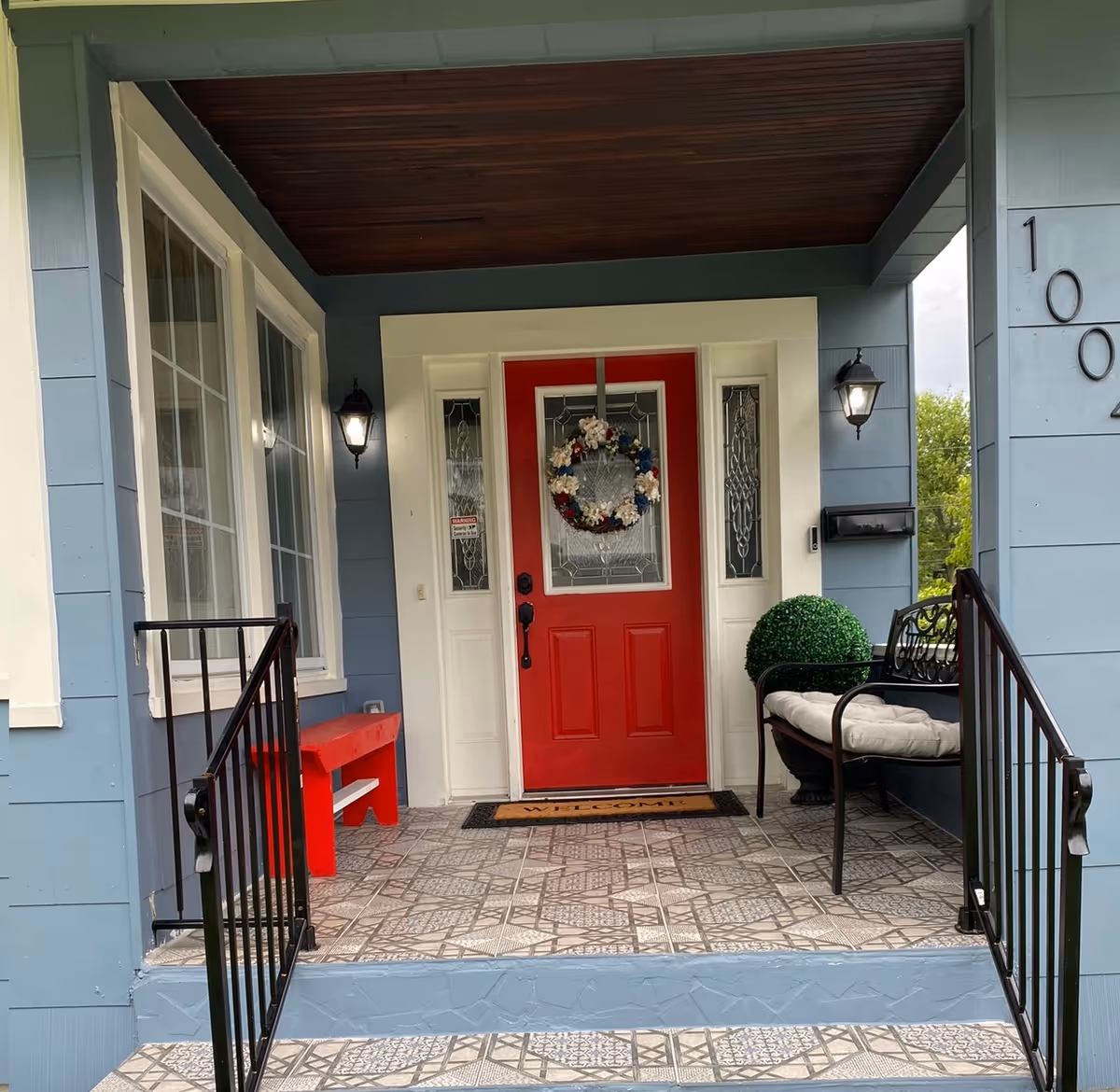 Front porch of a blue house with red door, wreath, bench, and patterned tile floor.