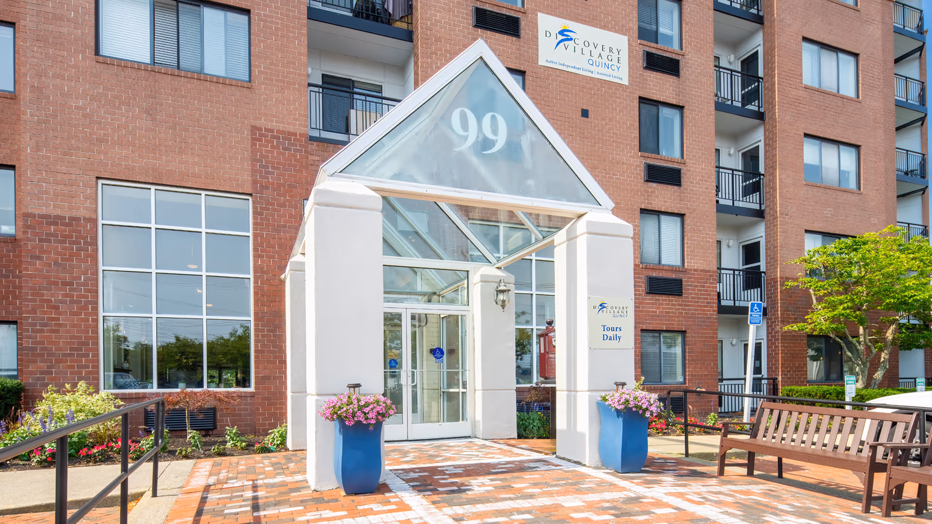 Brick senior living building with glass pyramidal entrance, benches, planters, and accessible walkway.