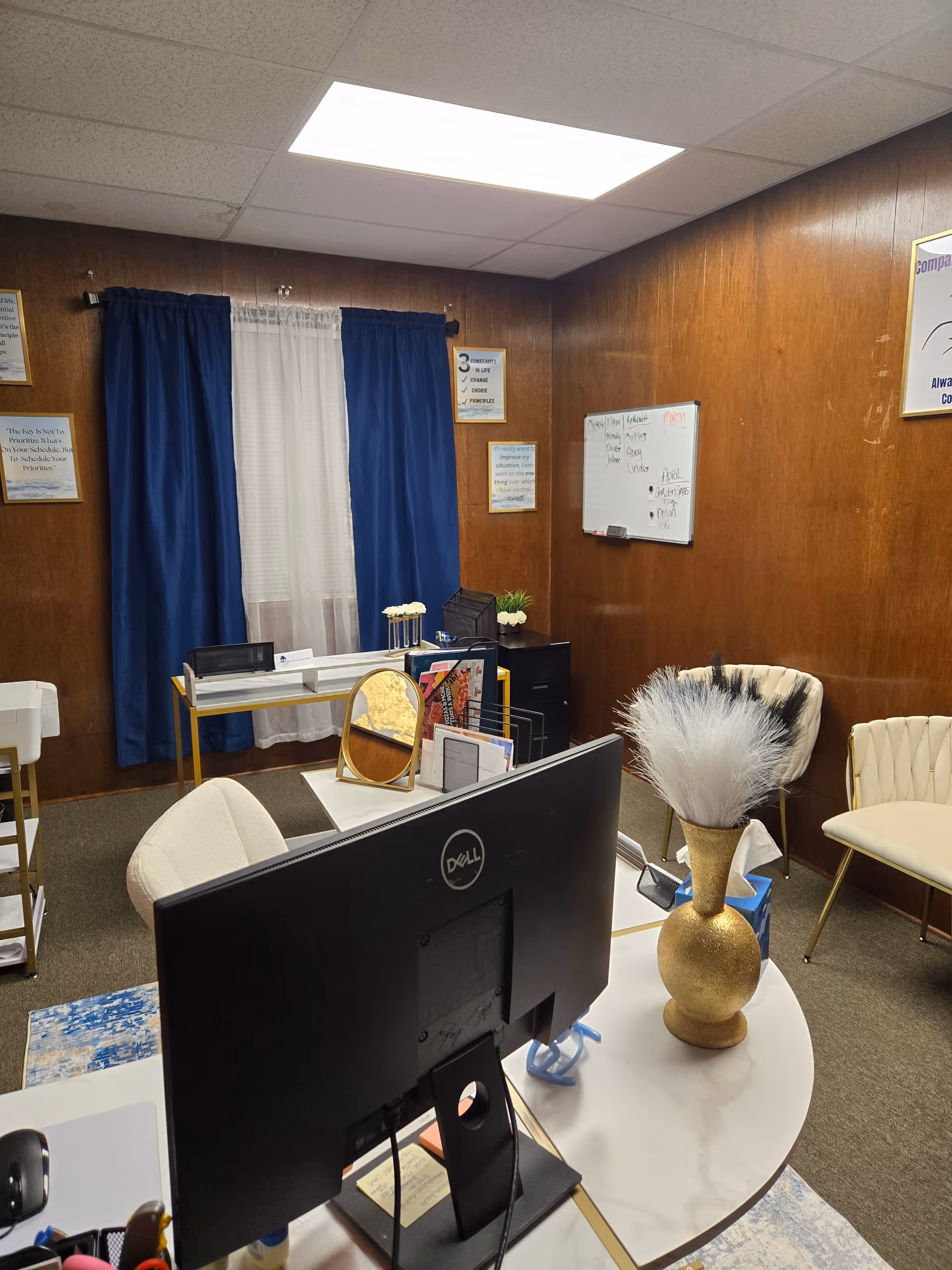 Reception-style office with a Dell monitor, whiteboard, blue curtains, and decorative items.
