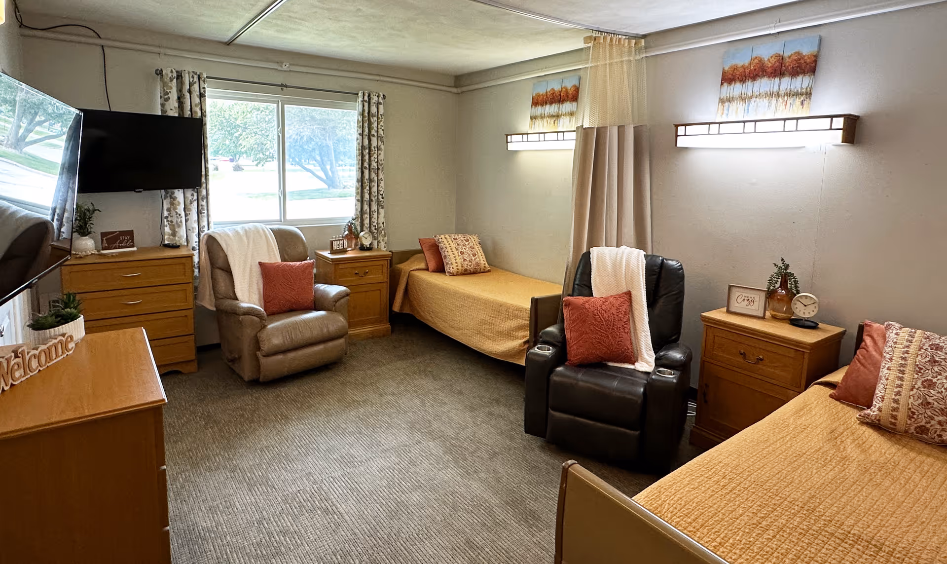 A cozy room with two single beds, each with a pillow and a quilted bedspread. Between the beds are two wooden nightstands with decorative items including a clock and framed signs. There are two comfortable recliner chairs with throw pillows and blankets. A window with floral curtains lets in natural light. A flat-screen TV is mounted on the wall above a wooden dresser with a welcome sign and small plant. The walls have modern light fixtures and artwork above the beds.