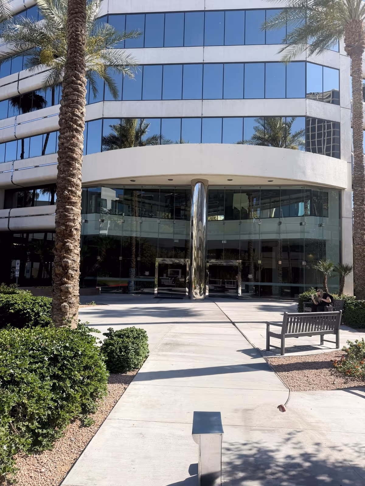 Glass-front building entrance with curved canopy, palm trees, and a bench.