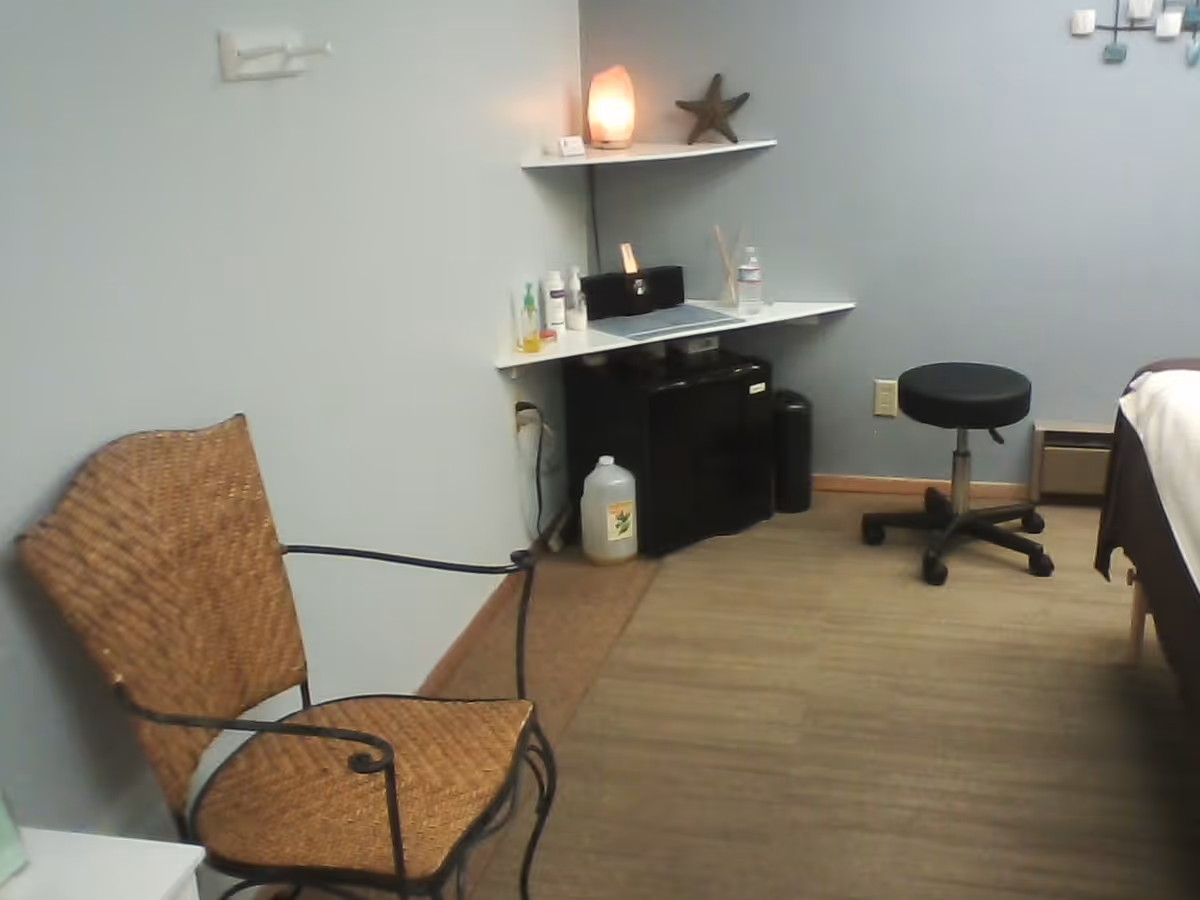 Interior treatment room with wicker chair, shelves, a bed, mini-fridge, and rolling stool.