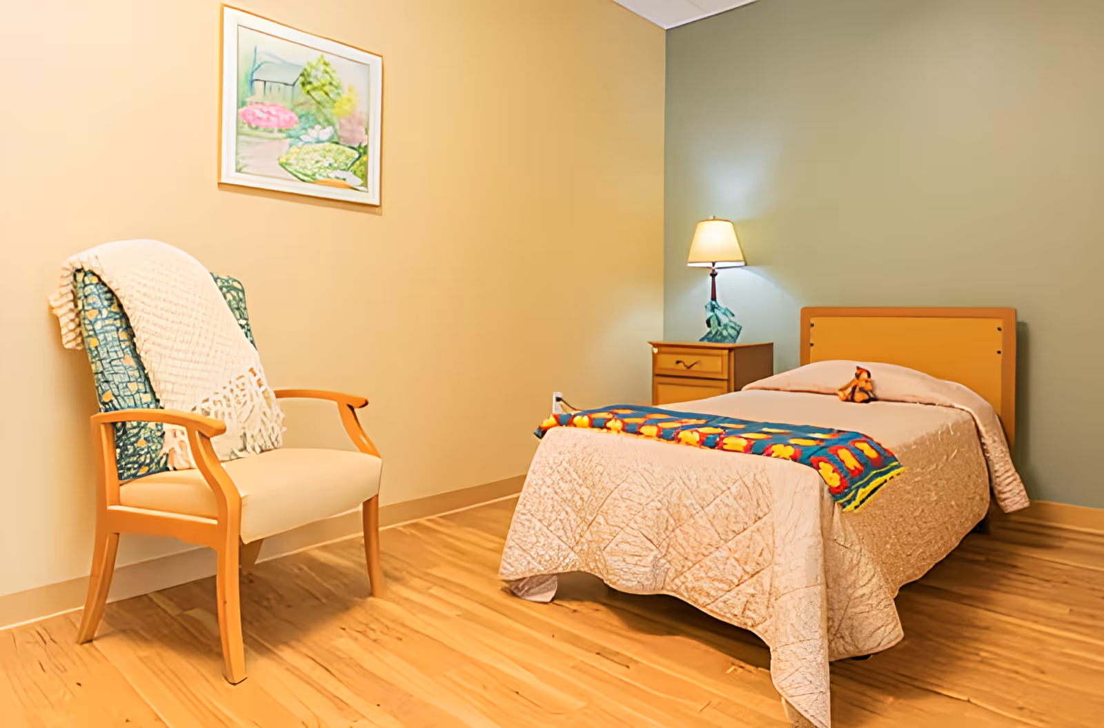 A simple bedroom with a single bed covered with a beige quilt and a colorful blanket at the foot. A small stuffed animal is placed on the bed. Next to the bed is a wooden nightstand with a lamp. On the left side of the room is a wooden armchair with a cushion and a white throw blanket draped over the back. A framed painting of a garden scene hangs on the wall above the chair. The floor is wooden, and the walls are painted in two different colors, beige and light green.
