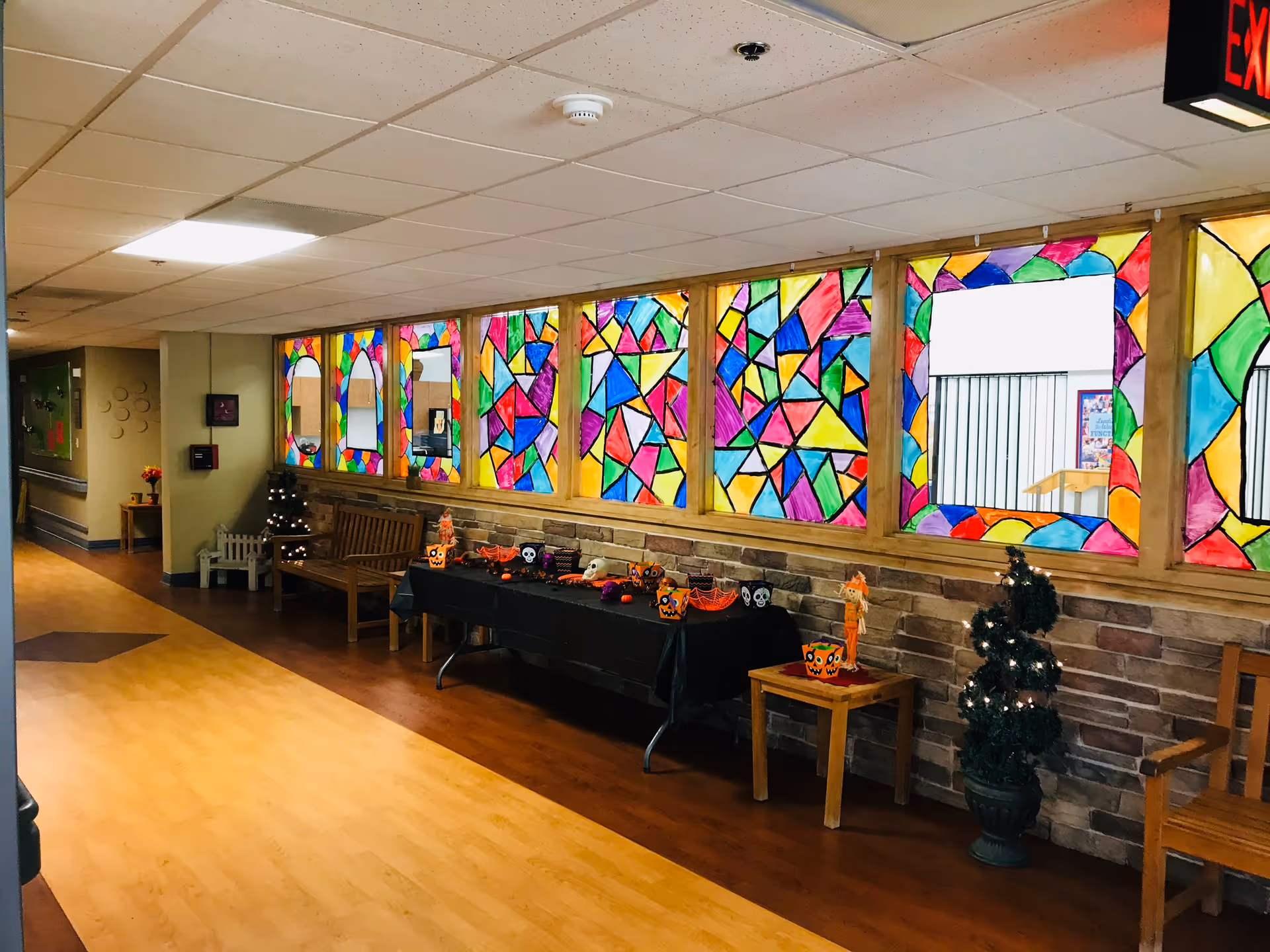 A hallway in a senior living facility with colorful stained glass-style window decorations. The hallway has wooden benches, a table covered with a black cloth decorated with Halloween-themed items, and small decorated Christmas trees. The floor is wooden, and the walls have a stone-like lower half with a light-colored upper half. An exit sign is visible on the ceiling.