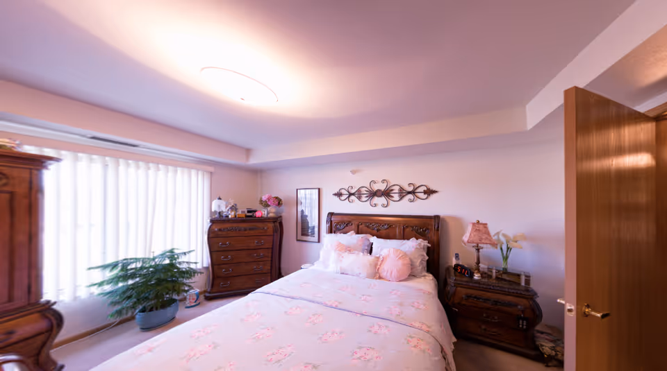 Cozy bedroom featuring a carved wooden bed with floral bedding, matching dressers and nightstands, a potted plant and vertical blinds.