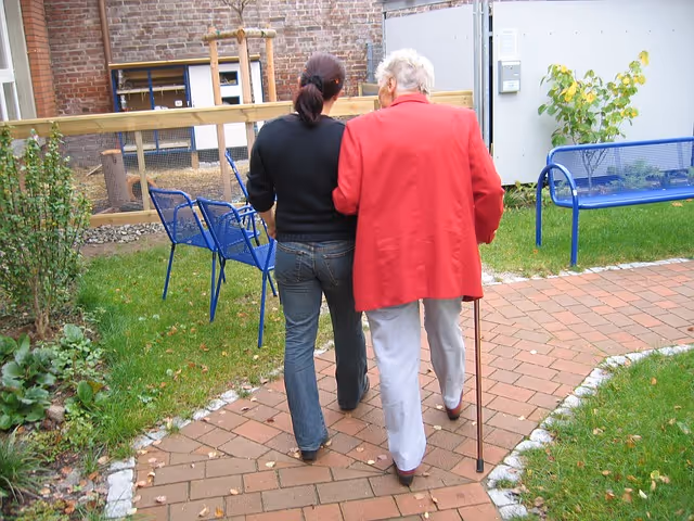 Two people walk along a brick path outdoors; one elderly person uses a cane.