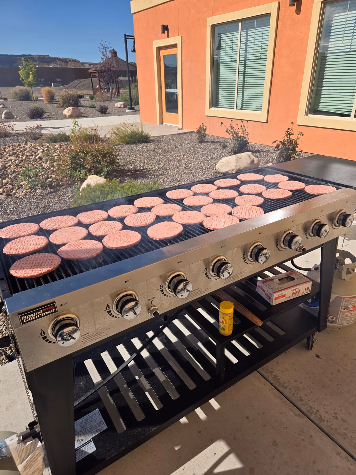 Outdoor grill loaded with hamburger patties beside an orange building.