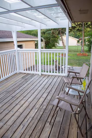 Wooden covered deck with white railings and outdoor chairs overlooking a green lawn.