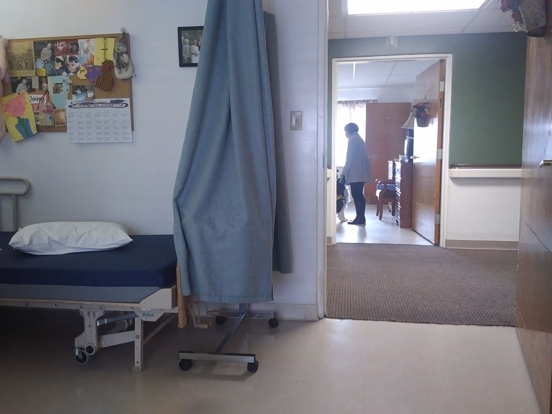 A hospital-style bedroom with a bed, privacy curtain, and a person standing in a doorway.