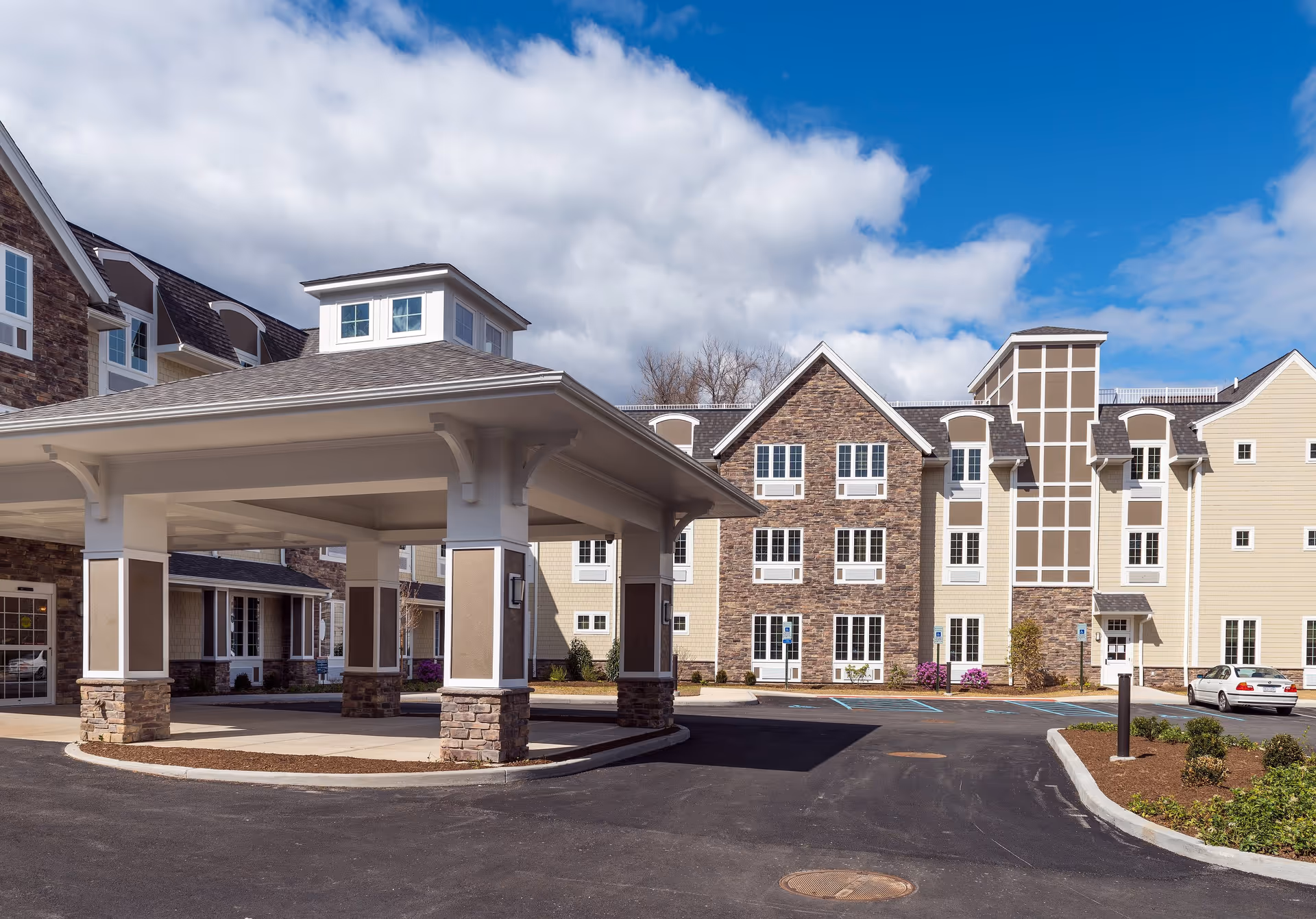 Exterior view of The Ambassador of Scarsdale senior living facility showing a covered entrance with stone pillars, a paved driveway, parking spaces including handicapped spots, and a multi-story building with stone and beige siding under a partly cloudy blue sky.