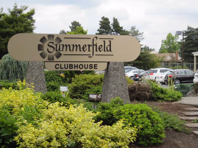 Entrance sign reading "Summerfield Clubhouse" surrounded by landscaping with parked cars in the background.