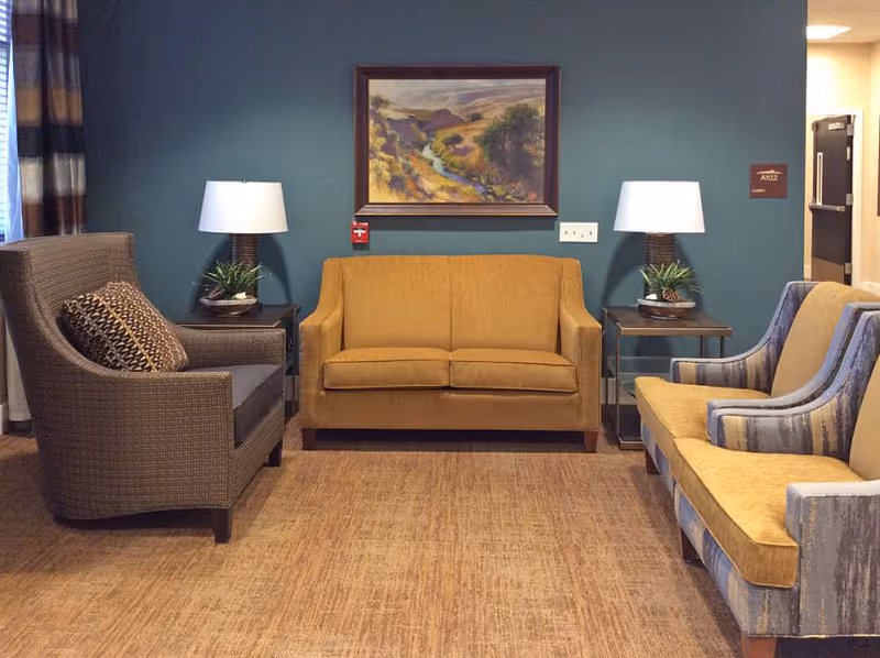 A cozy seating area in an assisted living facility with a mustard yellow loveseat in the center, a patterned armchair with a decorative pillow on the left, and a striped armchair with yellow cushions on the right. Two side tables with lamps and small plants flank the loveseat. A framed landscape painting hangs on the teal-colored wall behind the loveseat.