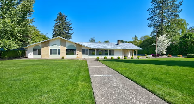 Single-story building with a light-colored exterior and a metal roof, surrounded by a well-maintained green lawn and trees under a clear blue sky. A concrete walkway leads up to the entrance of the building.