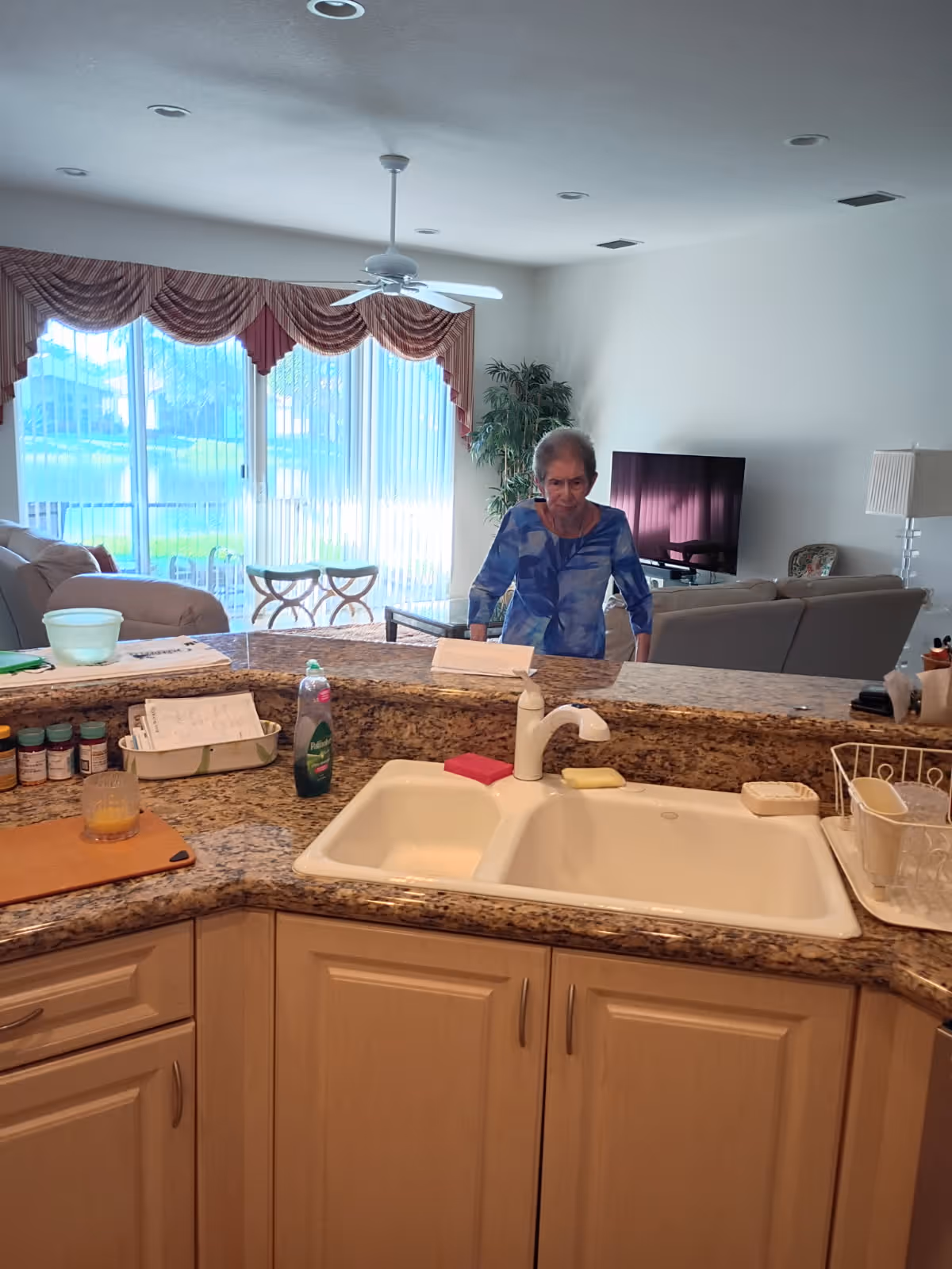 An elderly woman stands at a granite kitchen counter in an open living area.