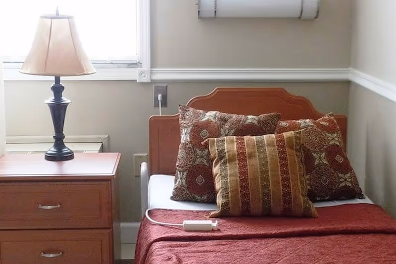 Single bed with patterned pillows and a bedside table with a lamp next to a window in a small bedroom.