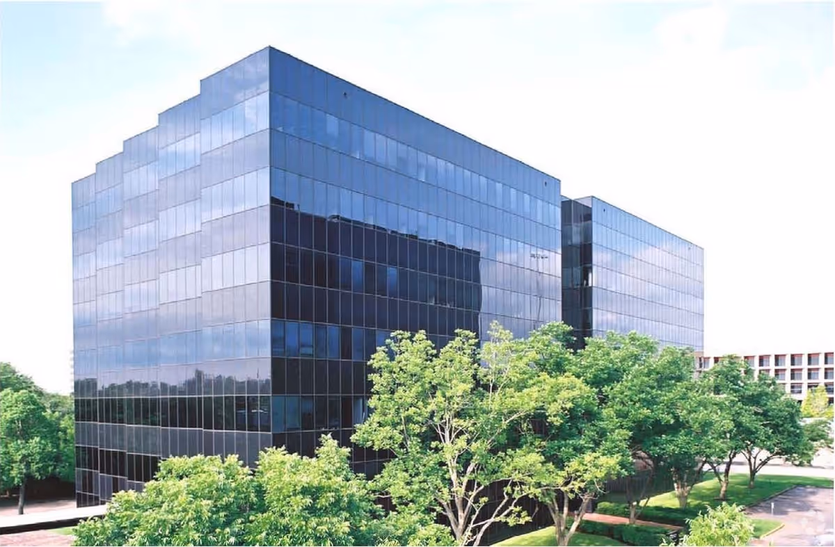 A modern glass building with surrounding green trees under a clear sky