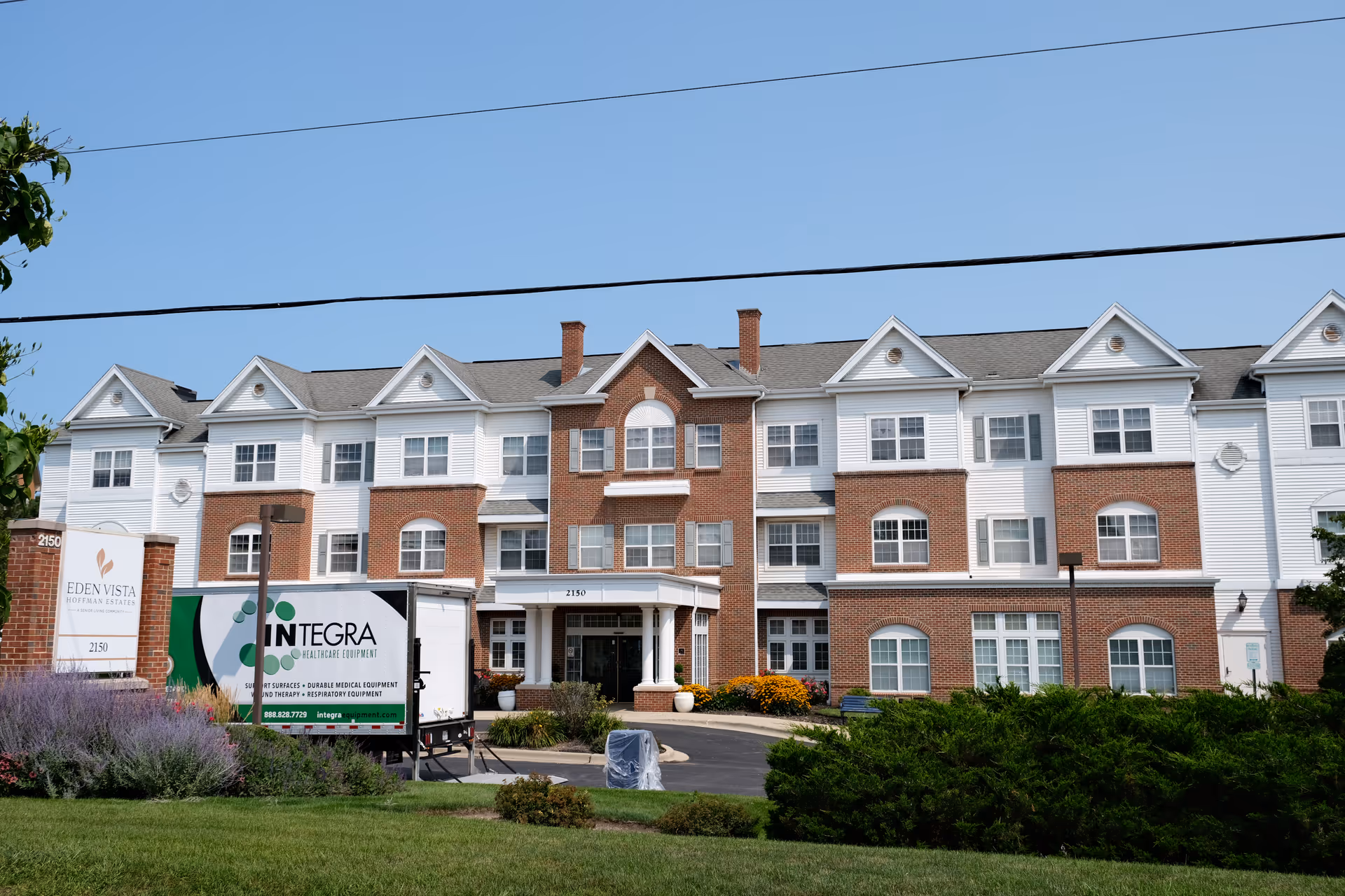 Front exterior of the Eden Vista Hoffman Estates three-story brick and white-siding senior living building with landscaped entrance and a parked delivery truck.