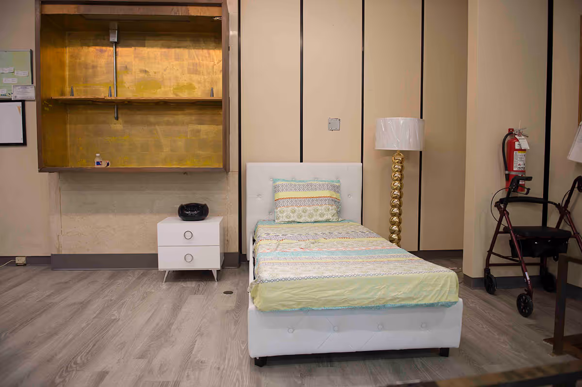 A bedroom interior featuring a white bed with a colorful striped blanket, a nightstand with a black object, a gold lamp, and a walker in the corner.
