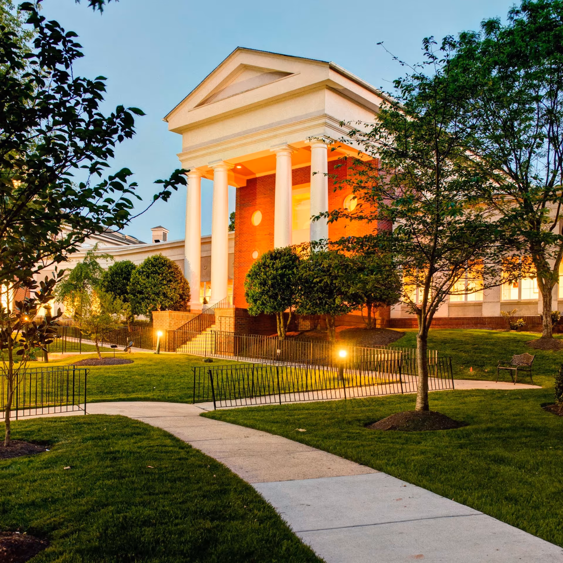 Exterior view of a stately building with large white columns and a triangular pediment, surrounded by well-maintained green lawns, trees, and a paved walkway, illuminated by warm lighting during dusk.