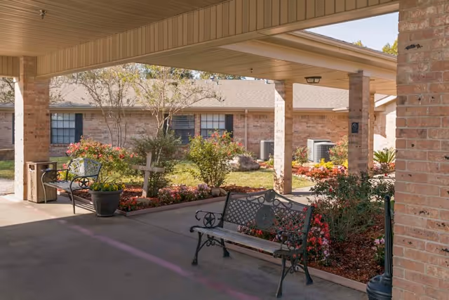 Covered entrance courtyard of a brick senior living facility with metal benches, potted plants, and landscaped flowerbeds.