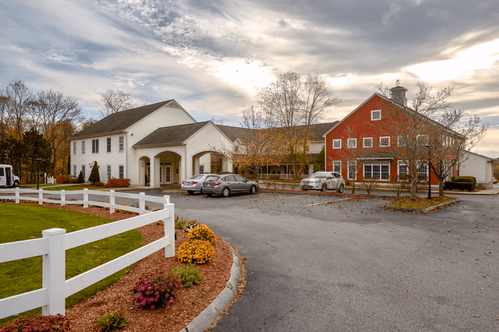 Exterior view of Benchmark at Billerica Crossings facility showing a two-story building with white and red sections, a parking lot with several cars, a white fence, landscaped flower beds, and trees with autumn foliage under a cloudy sky.